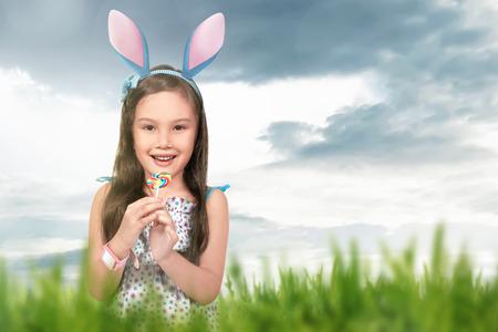 Asian little girl wearing bunny ears holding lollipop on green grass backgroundの写真素材