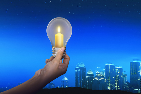 People hand holding candles light inside light bulb with modern city backgroundの写真素材