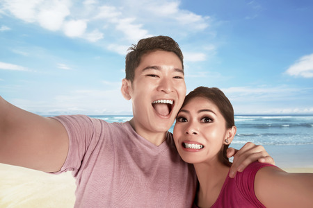 Asian couple in love making selfie photo with blue ocean as backdrop at beachの写真素材