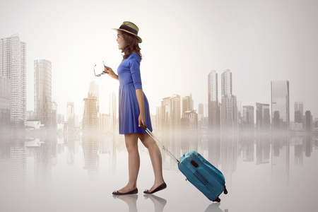 Young asian woman with hat holding suitcase over cityscape backgroundの写真素材