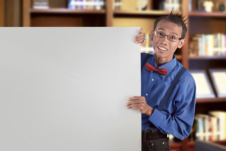 Happy asian nerdy man showing blank white board for copyspace on libraryの写真素材
