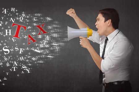 Young asian businessman using megaphone shouting notice TAX against wall backgroundの写真素材