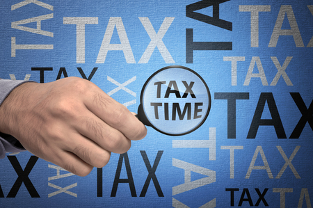 Business hand holding magnifying glass over TAX TIME text on blue backgroundの写真素材
