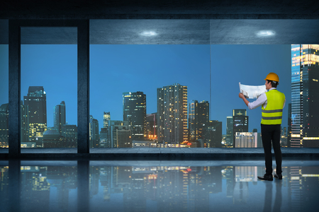 Young asian engineer man looking at his blueprints at room with cityscape backgroundの写真素材