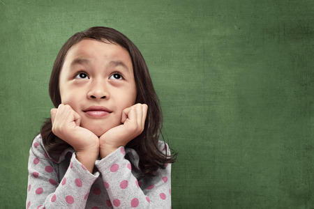 Beautiful asian little girl with hands in chin imagine something against chalkboard backgroundの写真素材