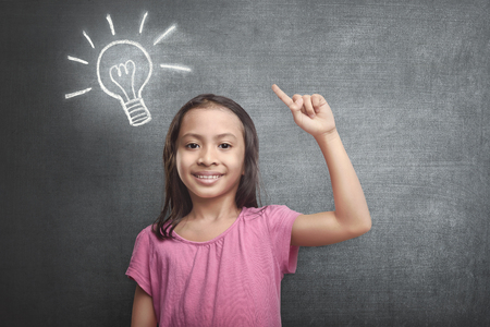 Cheerful asian kid with bright idea symbol above her head against blackboard backgroundの写真素材