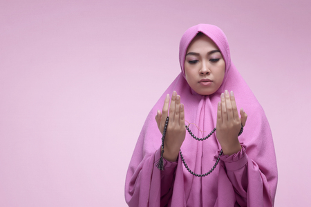 Young asian muslim woman praying with prayer beads over pink backgroundの写真素材