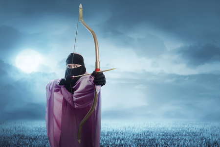 Young asian muslim woman in hijab ready to shoot an arrow in the nightの写真素材