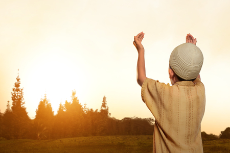 Back view of asian muslim kid with cap praying to god in nature during sunsetの写真素材