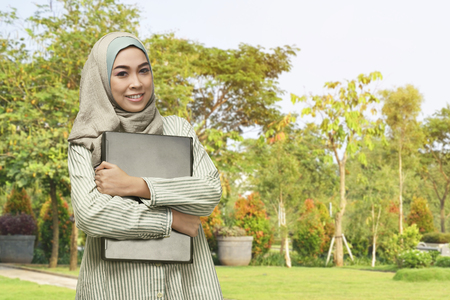 Beautiful asian muslim woman with smiley face holding laptop in the parkの写真素材
