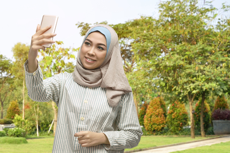 Beautiful asian muslim woman taking selfie photo with smartphone on the parkの写真素材