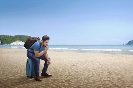Young asian man with backpack sitting on luggage on the beachの写真素材