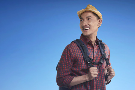 Happy asian backpacker wearing hat carrying backpack against bright backgroundの写真素材