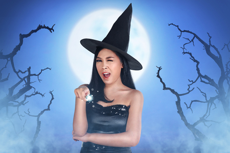 Smiling asian woman with witch dress holding spell wand at moonlightの写真素材