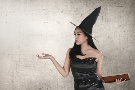 Young asian witch woman with hat holding spell book against grunge backgroundの写真素材