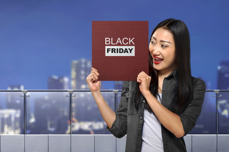 Happy asian woman holding board with Black Friday message over cityscape backgroundの写真素材