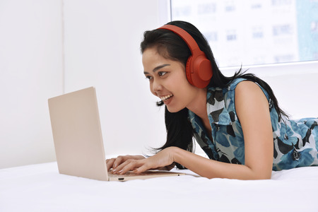 Happy asian woman using laptop and enjoying the music while lying on bedroomの写真素材