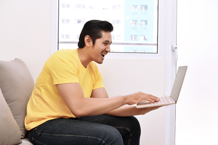 Happy asian man with laptop sitting on the couch at homeの写真素材