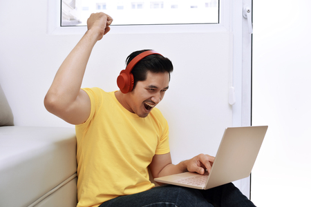 Happy asian man sitting with laptop and headphones in the living roomの写真素材