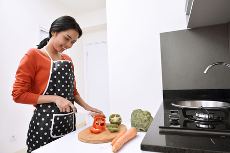 Smiling asian woman using knife to slice peppers and cooking in the kitchenの写真素材