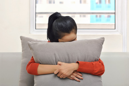 Young asian woman hugging and hiding behind a pillow in the sofaの写真素材