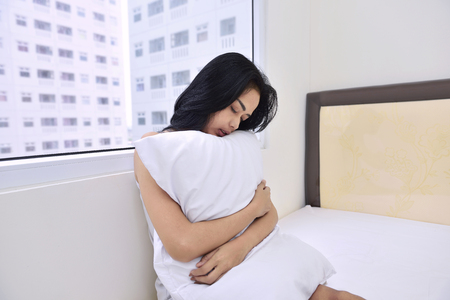 Beautiful asian woman sitting and hugging white pillow with window backgroundの写真素材
