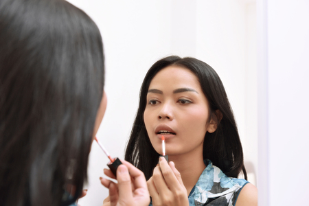Beautiful asian girl applying lipstick while looking on the mirror at homeの写真素材