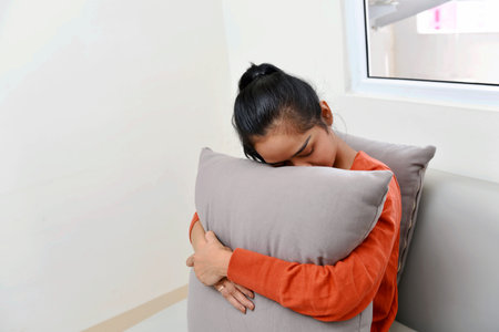Sad asian woman hugging a pillow and sitting on sofa at homeの写真素材