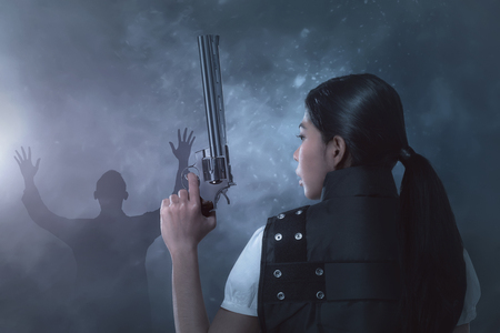 Young asian woman in a police uniform with pistol on her hands on dark backgroundの写真素材
