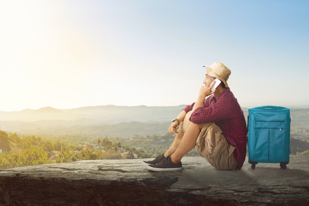 Young asian tourist with mobile phone relaxing on top of the rock over sky backgroundの写真素材