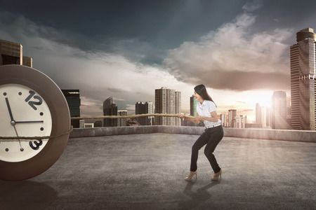 Young asian businesswoman using rope to pulling a clock with skyscrapers backgroundの写真素材
