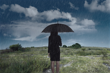 Businesswoman standing with an umbrella in rain over dark sky backgroundの写真素材