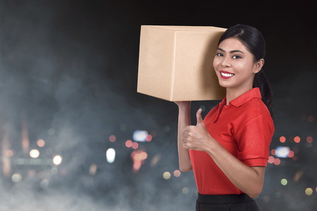 Beautiful asian female courier holding the package with blur lights backgroundの写真素材