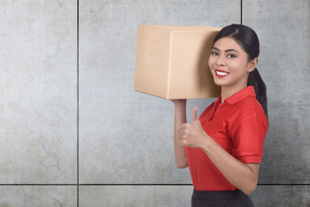 Smiling asian courier woman bring the parcel against gray wall backgroundの写真素材