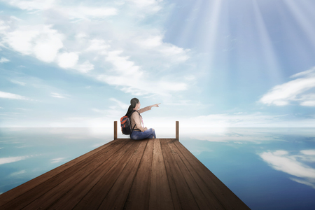 Young asian backpacker woman sitting and pointing blue sky at pierの写真素材