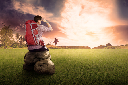 Rear view of asian traveler man with backpack sitting and looking at sunset view on the fieldの写真素材