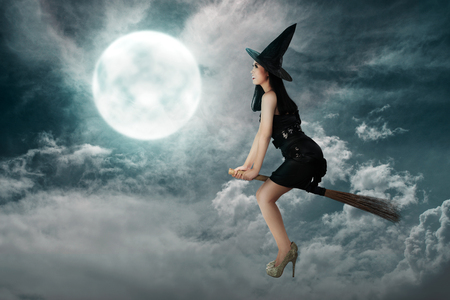 Happy asian witch woman flying above a broomstick with full moon backgroundの写真素材