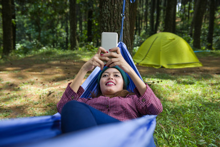 Smiling asian woman with phone resting in hammock on the forestの写真素材
