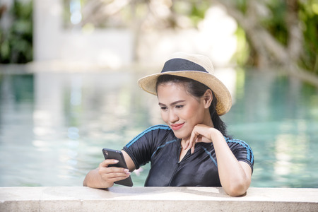 Smiling asian woman with straw hat using mobile phone on the poolsideの写真素材