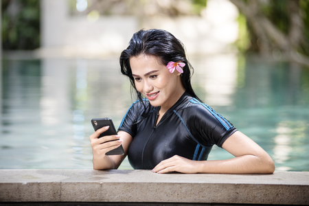 Happy asian woman with swimsuit using mobile phone in the poolsideの写真素材