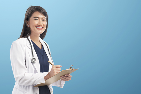 Pretty asian doctor woman with lab coat writing notes on the clipboard over blue backgroundの写真素材