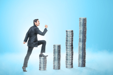 Attractive asian businessman going to the top of coins stack over blue backgroundの写真素材