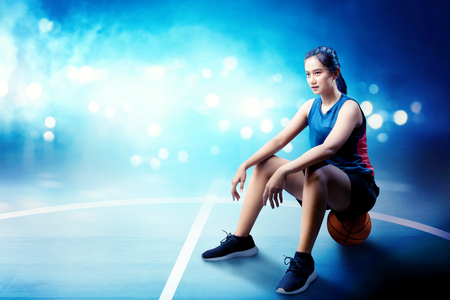 Smiling asian basketball player woman sitting on the ball in basketball courtの写真素材