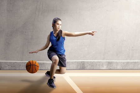 Beautiful asian woman in sport jersey dribbling the basketball with left arm pointing in the basketball court with wooden floor and concrete wall backgroundの写真素材