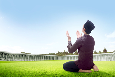 Rear view of asian man sitting in praying position on grass raise the hands and gazing the sky on the outside the mosqueの写真素材