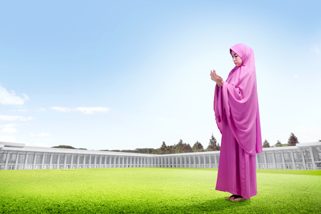 Beautiful asian woman in pink veil standing on grass raise the hands and look down outside the mosqueの写真素材