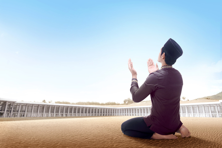 Back view of asian man sitting in praying position on desert raise the hands and gazing the sky on the outside the mosqueの写真素材