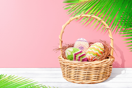 Colorful easter eggs on the nest in wooden basket on white wooden table with palm leaves and pink wall background. Happy Easterの写真素材