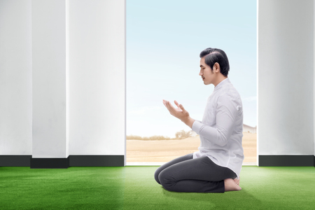 Handsome asian man sitting in praying position close his eyes and raise the hands on the carpet inside the room with open door and blue sky backgroundの写真素材