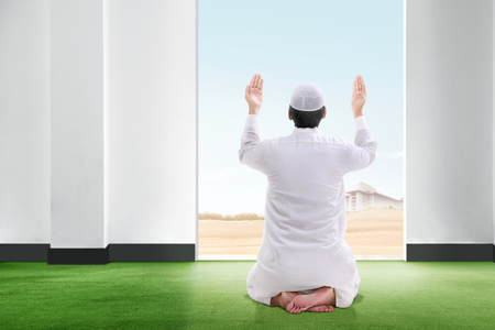 Back view of asian muslim man sitting in praying position on the carpet raise the hand and gazing the sky from inside the room with open door and blue sky backgroundの写真素材